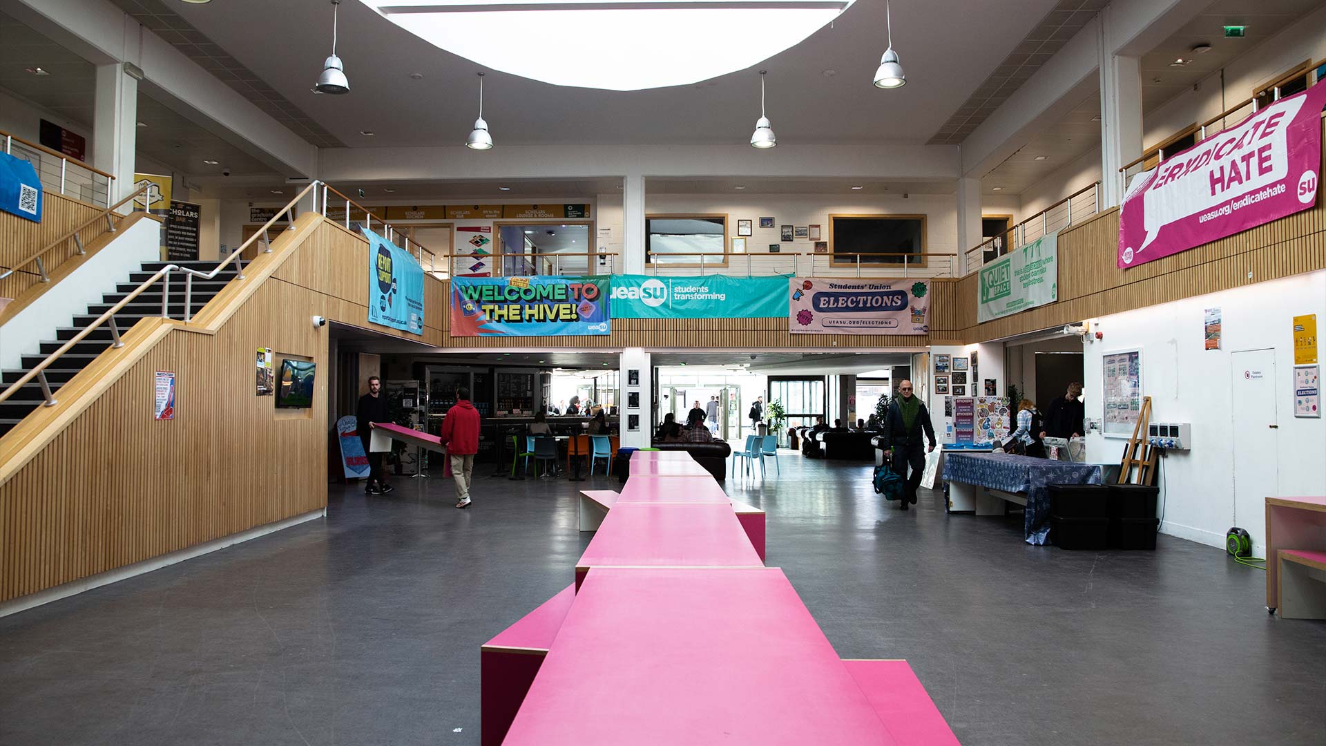 Open landscape shot of the Hive. Long pink tables run down the centre of the image, to the top you can see various advertisement posters.